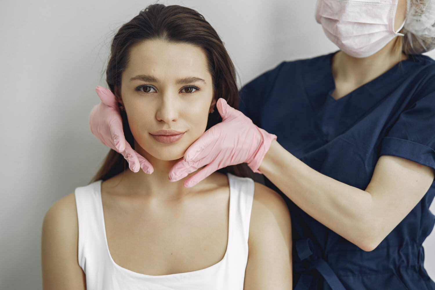 Profissional com luvas rosas avaliando o rosto de uma paciente. Representação de consulta de pós-operatório de bichectomia em Feira de Santana.