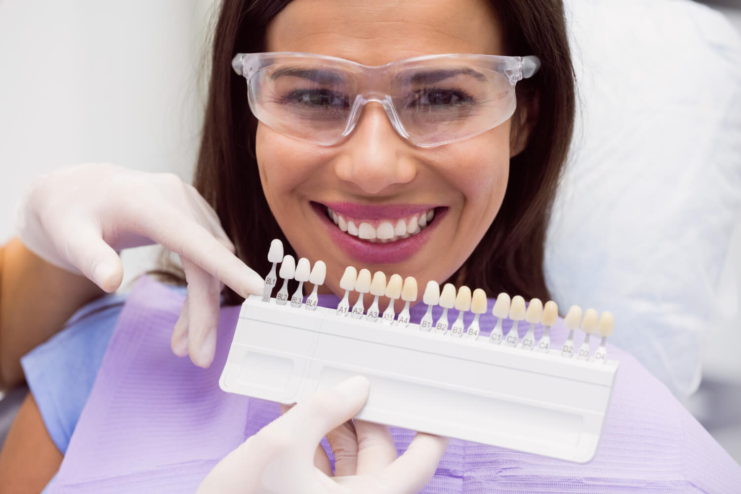 Mulher sorridente com óculos de proteção enquanto dentista segura escala de cores para lentes de contato dental na Clínica Dra. Camila Freitas em Feira de Santana.