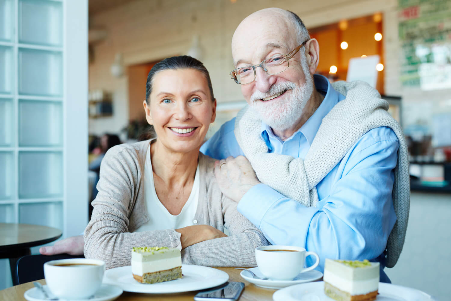 Casal idoso sorridente em um café, celebrando a reabilitação oral com implante dentário e prótese protocolo na Clínica Dra. Camila Freitas em Feira de Santana.